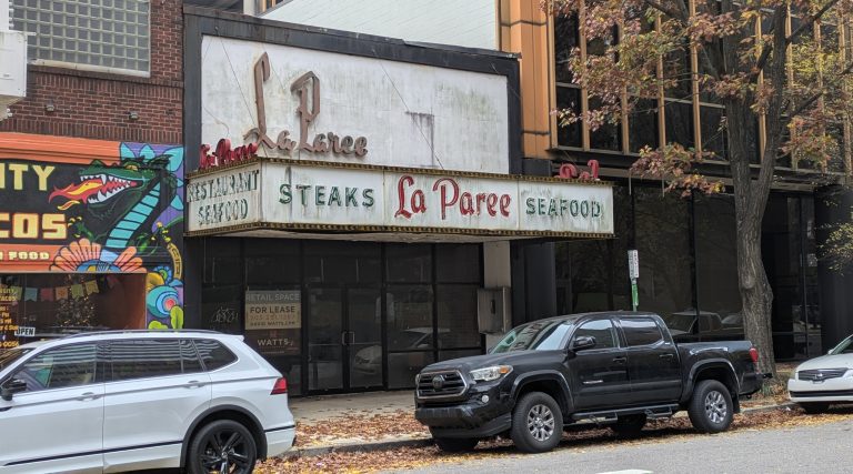 Image of historic restaurant space in downtown Birmingham, including a painted sign with neon highlights. There are two cars in front of the building.