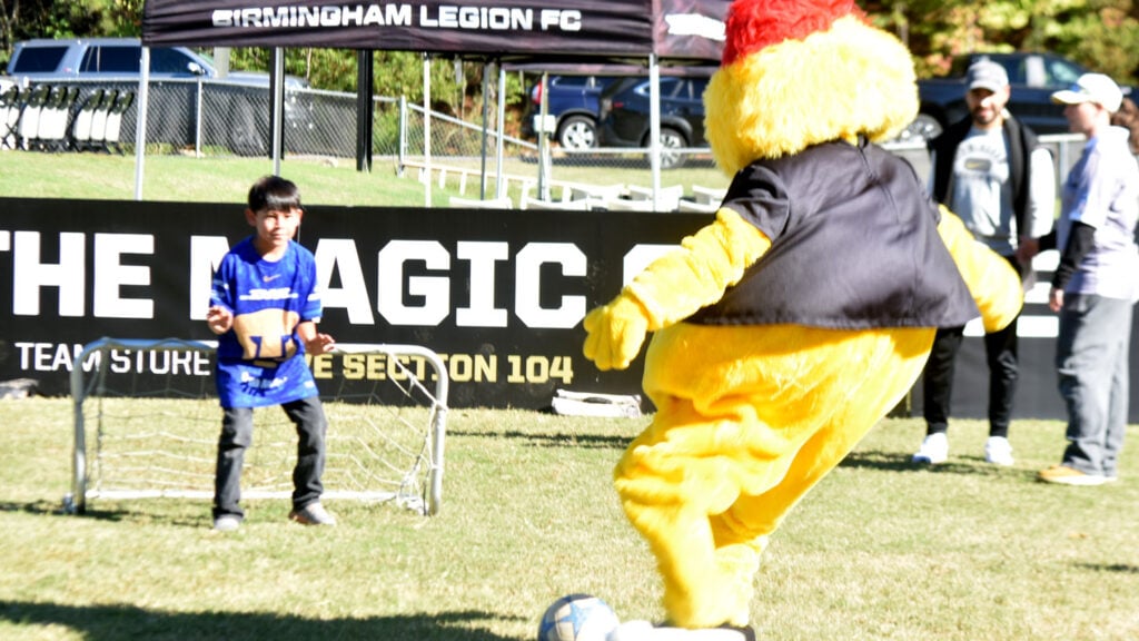 Noigel, a furry yellow mascot for the Birmingham Legion FC, scores a goal against a young child wearing a blue shirt and blue jeans on the playing field.